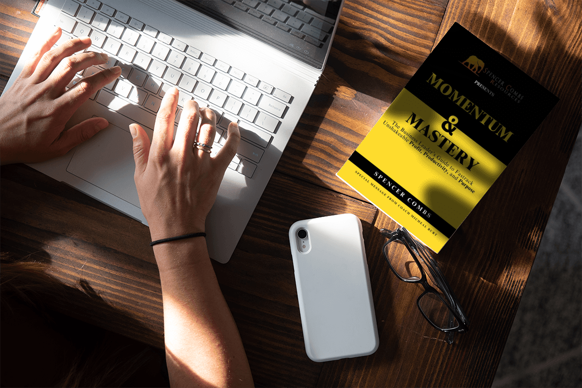 Momentum & Mastery book by Spencer Combs on a workspace desk next to a laptop, phone, and reading glasses