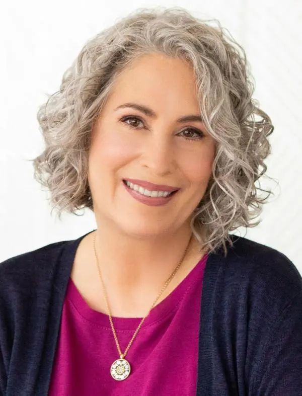 Portrait of a smiling woman with curly gray hair wearing a pink blouse and a pendant necklace