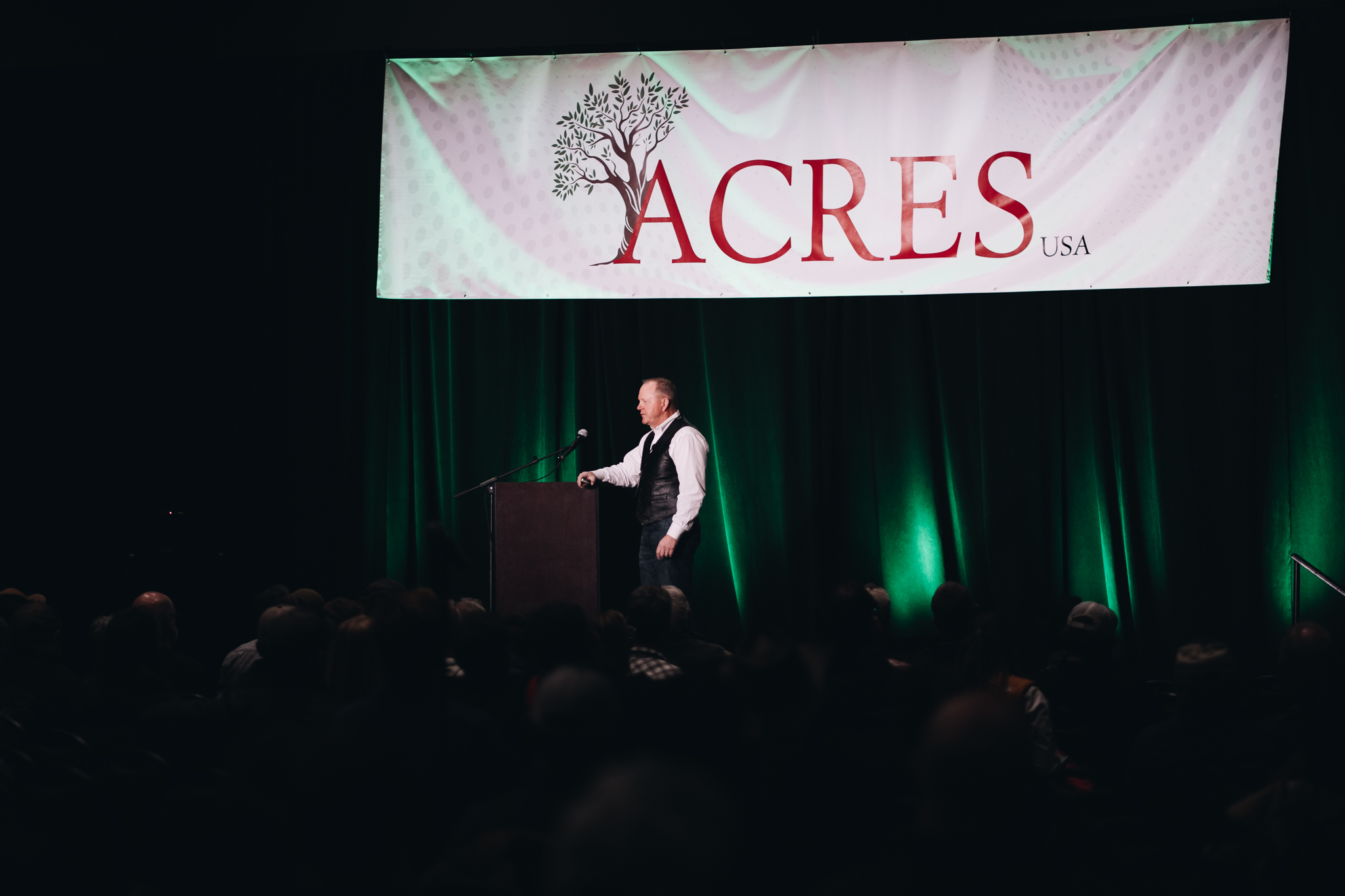 Keynote speaker presenting on stage at the Eco-Ag Conference under an Acres U.S.A. banner