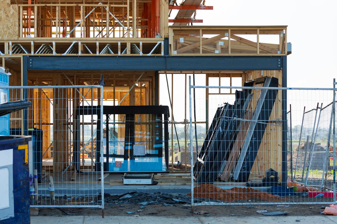Two-storey timber-framed house under construction behind temporary mesh site fencing, with stacked lumber and building materials.