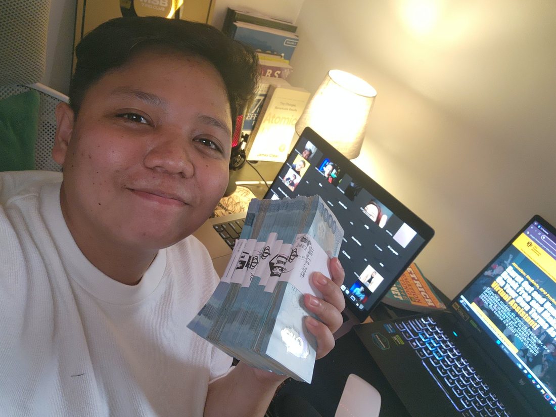 Bhryx smiling while holding a thick stack of ₱1,000 bills at a desk with laptops