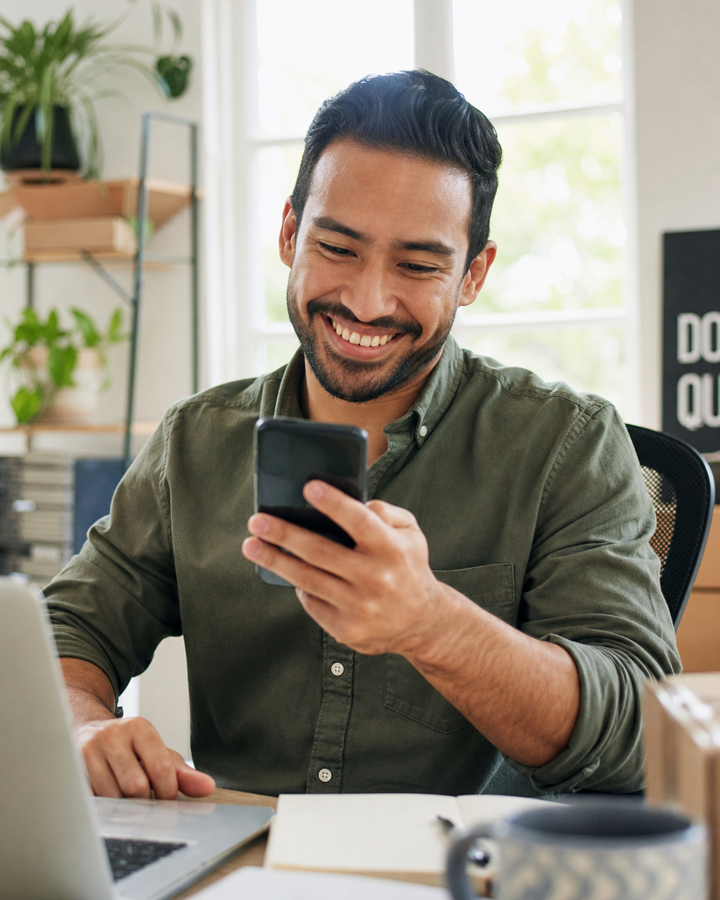 Latino male small business owner smiling as he reads a positive email reply on his phone at a bright sunlit home office desk
