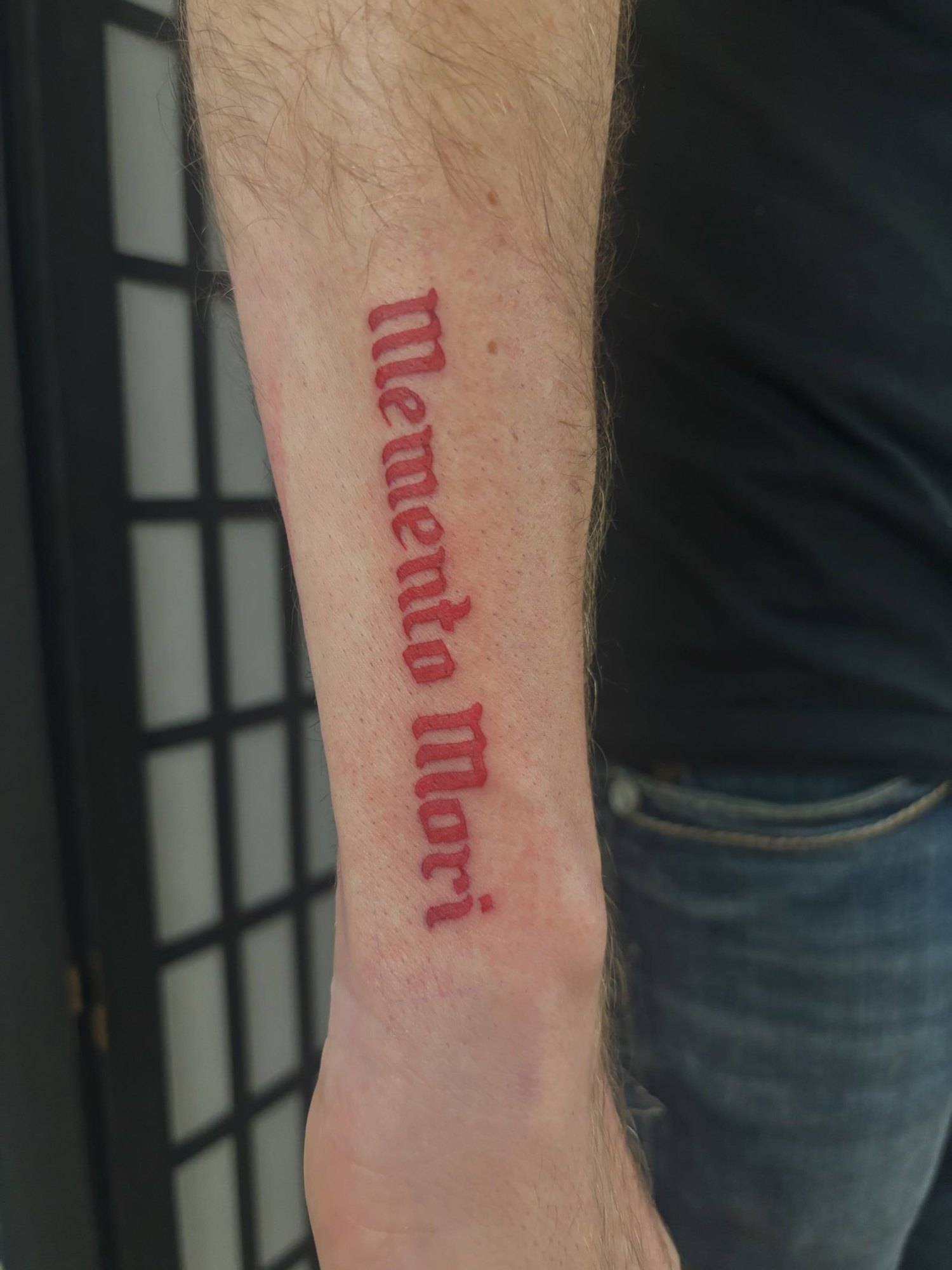 Memento Mori in red blackletter script down the forearm, by Carmelo