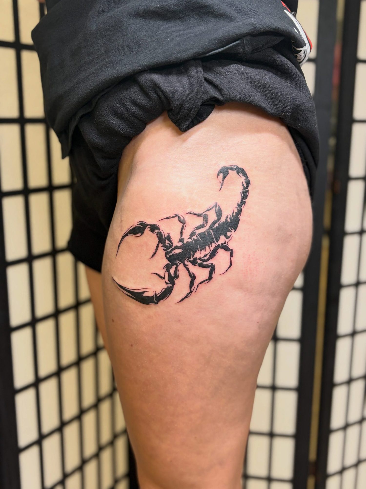 Black scorpion with its tail curled over its back on the outer thigh, by Peyton
