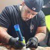 Gutta Banks in a cap and black gloves, tattooing