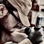 Carmelo in a cap and glasses, leaning in to tattoo