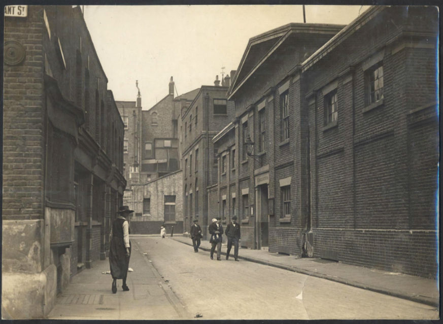 A historic black-and-white photograph of Mint Street Workhouse in Southwark, showing a long, plain brick building lining a narrow street. The building has simple, repetitive windows and a slightly worn appearance. In the foreground, a lone figure walks along the pavement, adding a sense of scale and quietness to the scene. The street is largely empty, with minimal traffic or activity, giving the image a stark, institutional feel typical of early 20th-century urban environments.