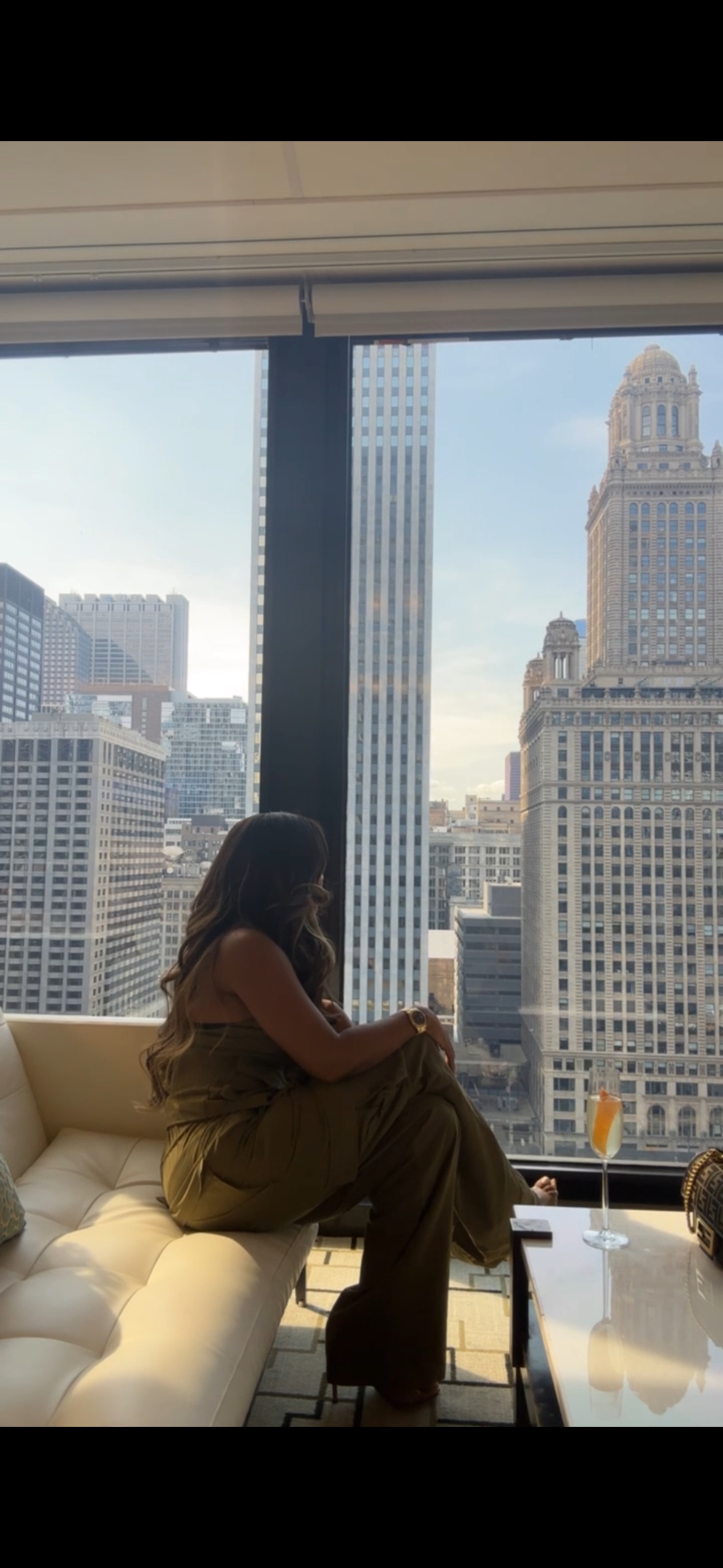 A beauty business owner sitting by a floor-to-ceiling window overlooking a city skyline, mid-afternoon, glass of champagne on the table beside her.