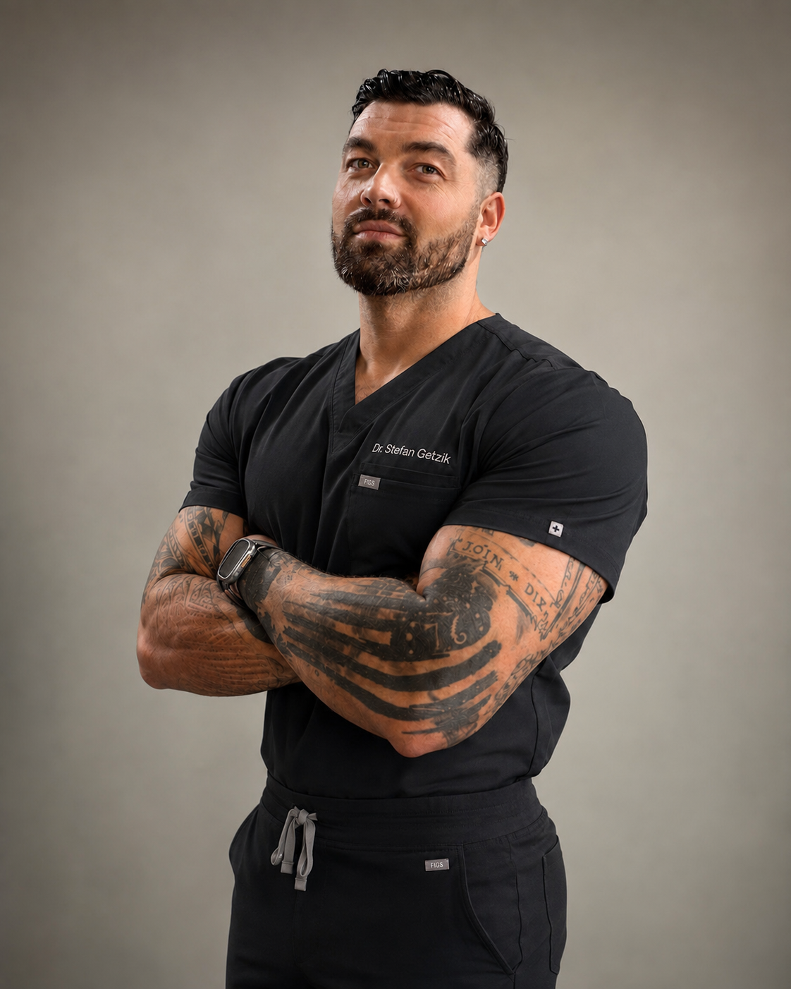 Dr. Stefan M. Getzik, DC, in black scrubs with his name embroidered on the chest, arms crossed in front of a warm grey backdrop