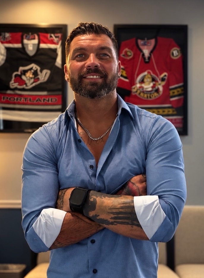 Dr. Stefan M. Getzik, DC, in black scrubs with his name embroidered on the chest, arms crossed in front of a warm grey backdrop