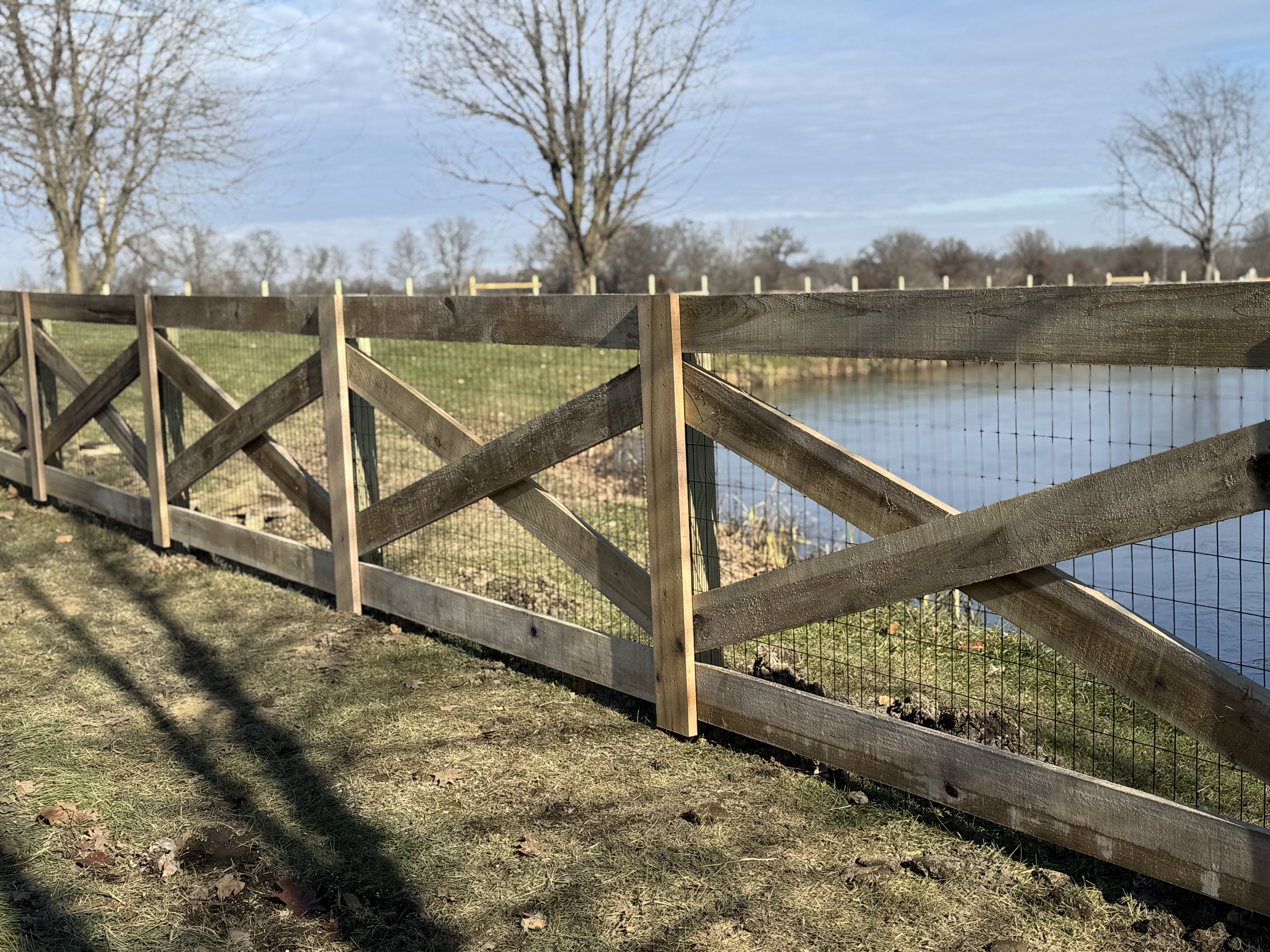 Rustic wood cross-buck split-rail fence with wire mesh backing near water in Granville, OH.