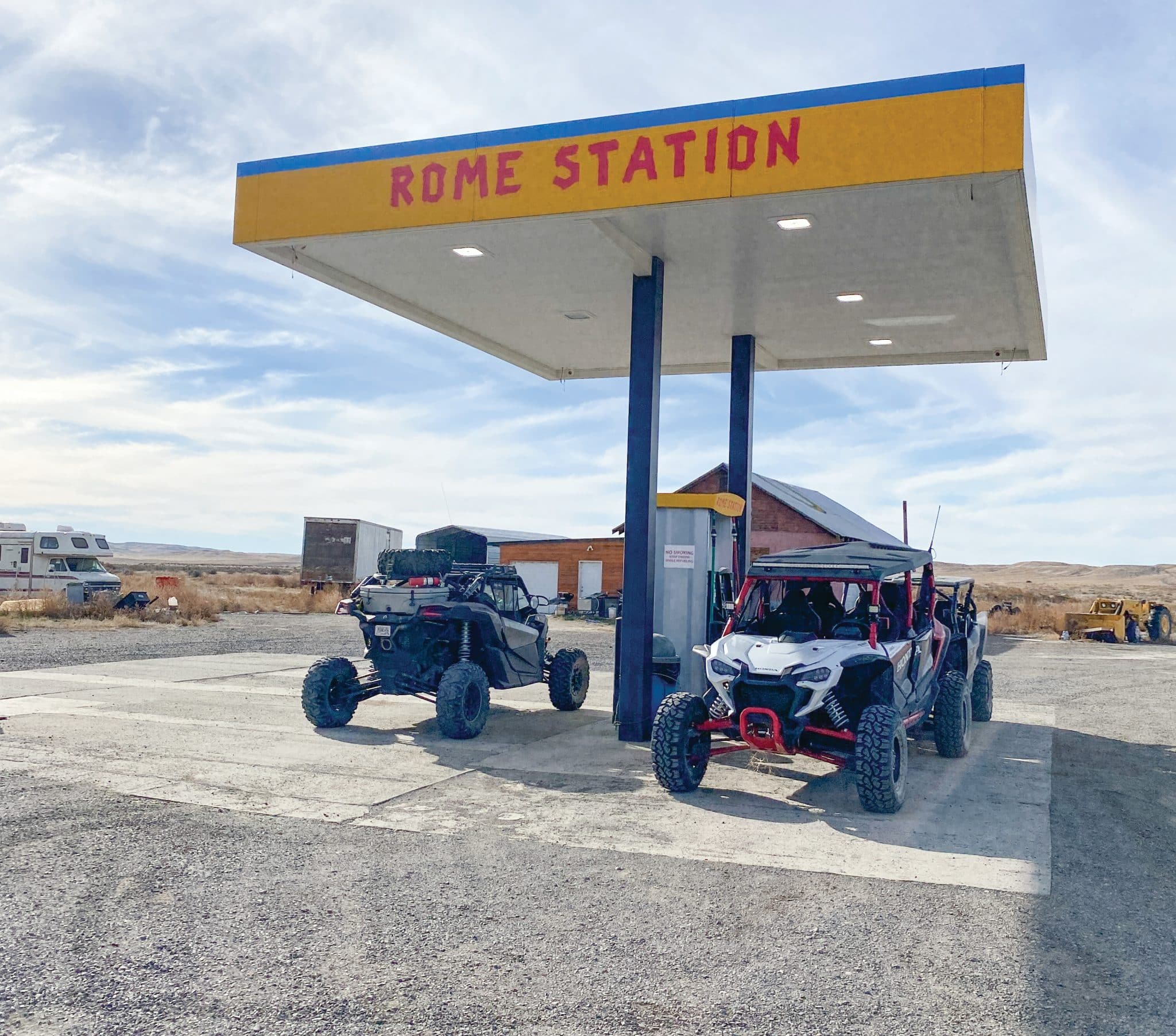 Two off-road UTV parked under a gas station canopy labeled "ROME STATION" in a remote desert area, with a small building and equipment visible in the background under a partly cloudy sky.