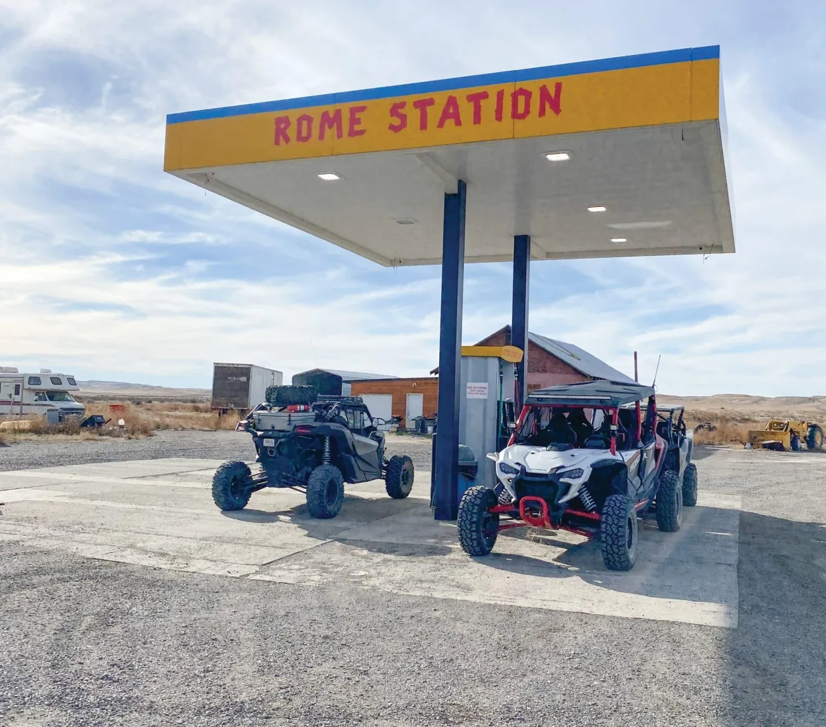 Two off-road vehicles with large tires are parked under a canopy at a "Rome Station" fuel stop in a rural area and ready for UTV registration. 