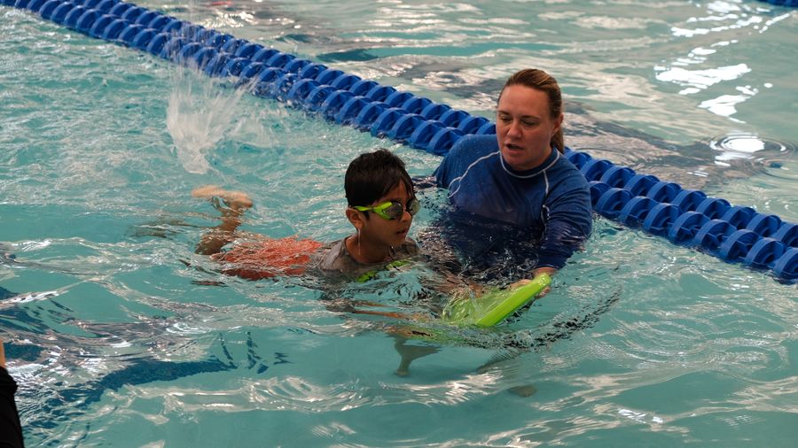 Instructor supporting a young swimmer with goggles on a kickboard beside a lane line