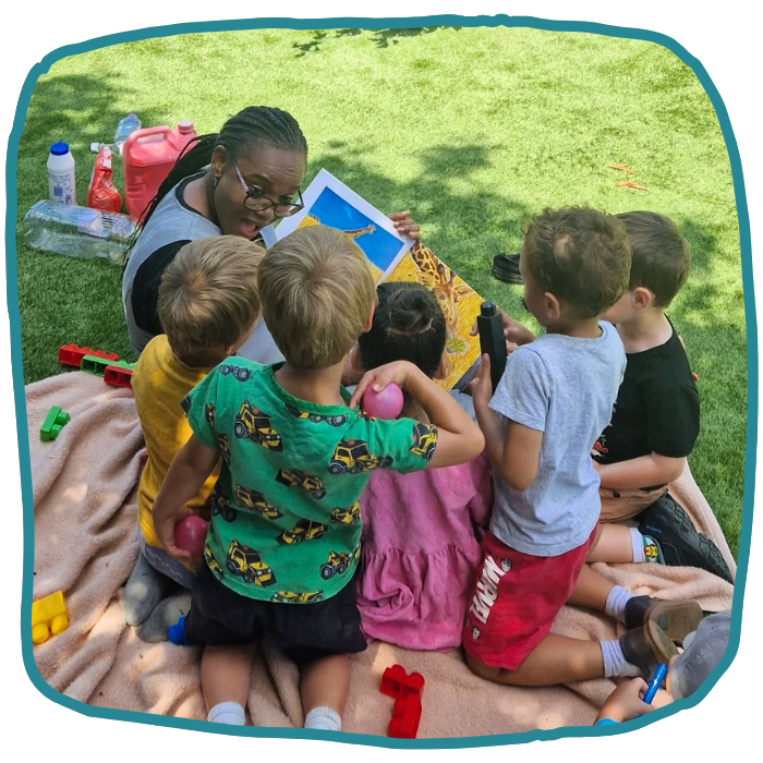 A woman shows a colorful picture book to a group of young children gathered around her on a blanket in a grassy park.