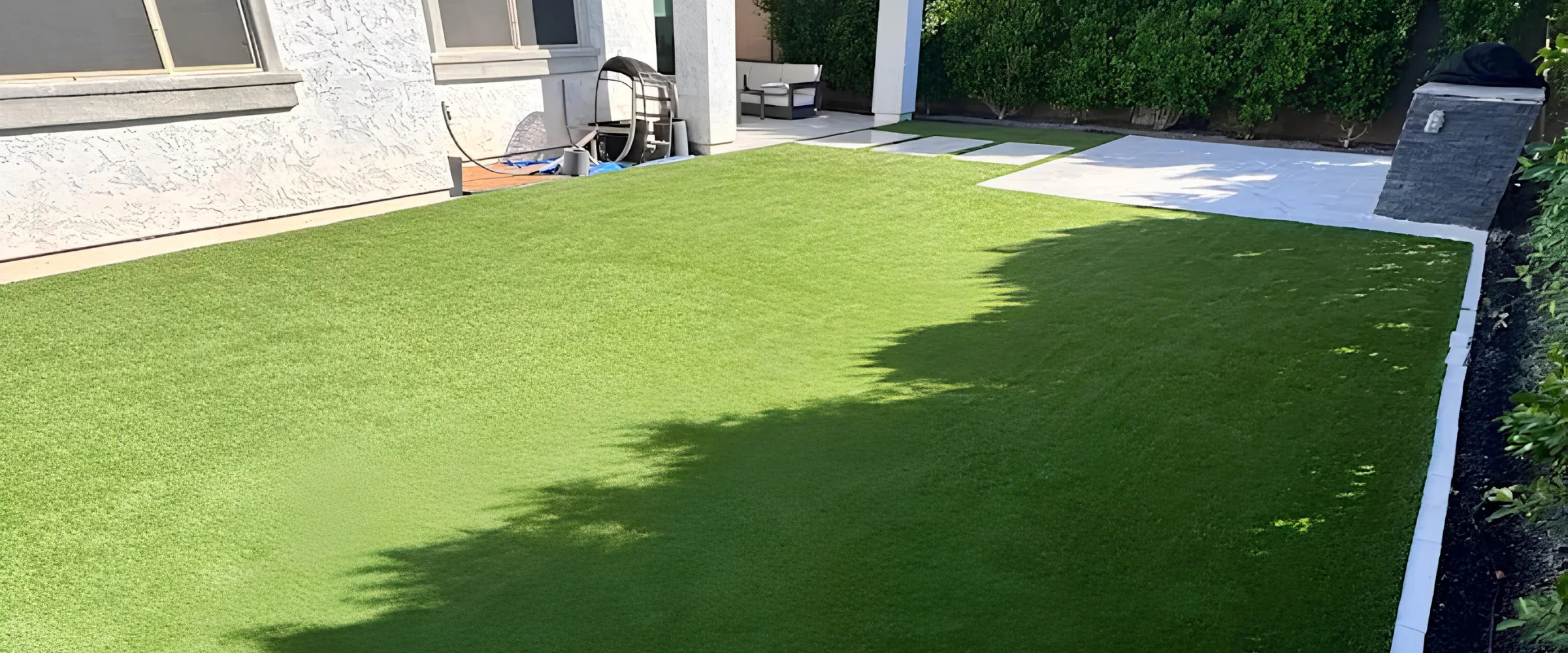 A clean backyard featuring artificial green turf, with a white stucco house, patio, and lush bushes in the background. A clean backyard featuring artificial green turf, with a white stucco house, patio, and lush bushes in the background.