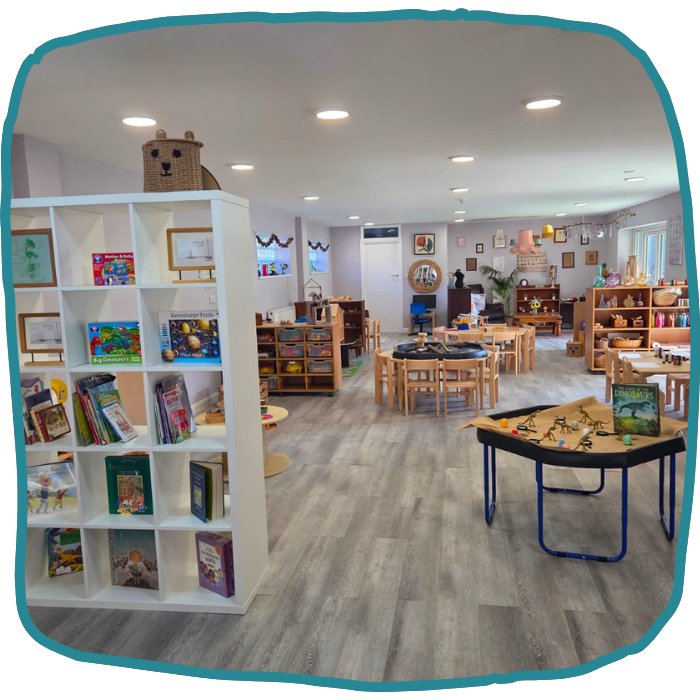A bright children’s classroom has picture books on white shelves, small tables and chairs, toy storage, and a play table with scattered toys at nursery in Croydon