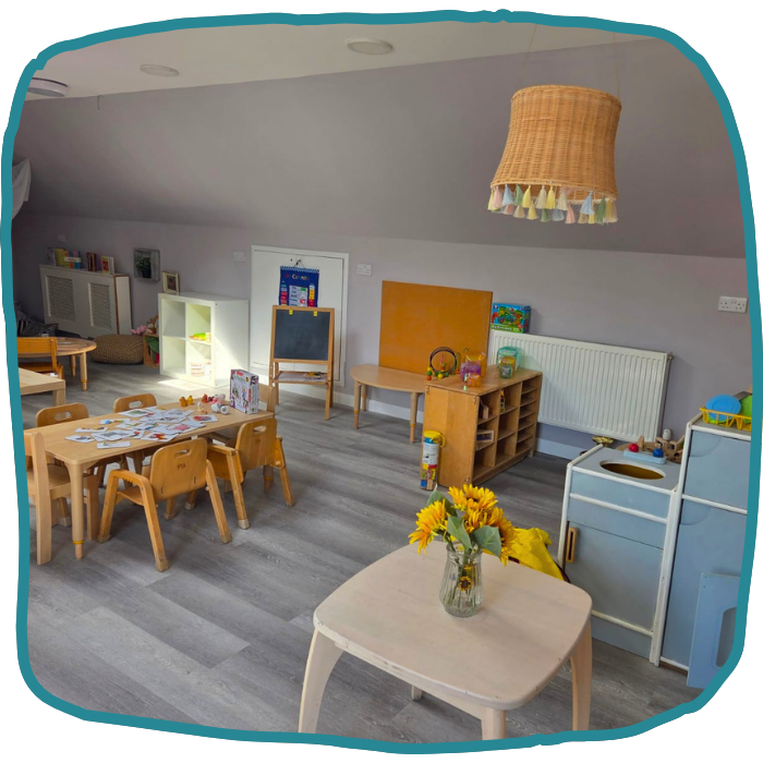 A bright classroom at Mini Treasures Croydon has child-sized tables and chairs, shelves, a chalkboard, and a foreground table with sunflowers.