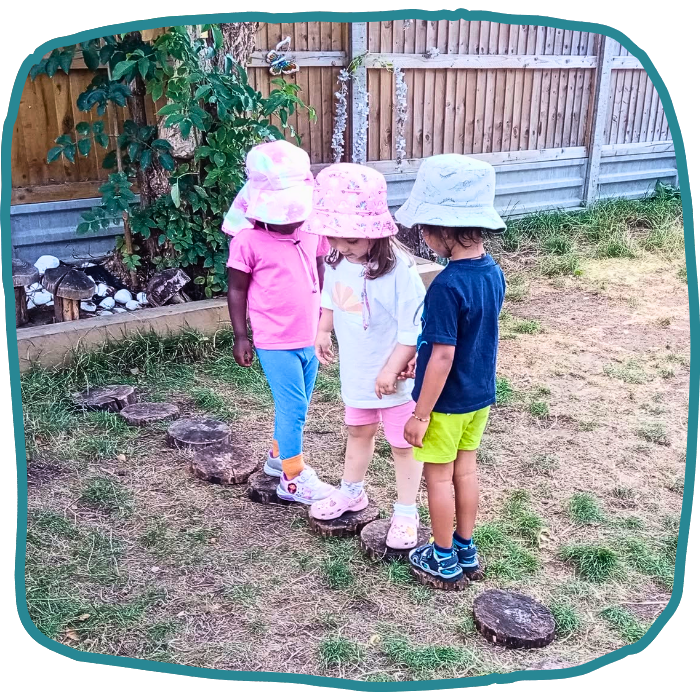 Three young children wearing sun hats stand together on stepping-stump rounds in a grassy backyard beside a wooden fence.