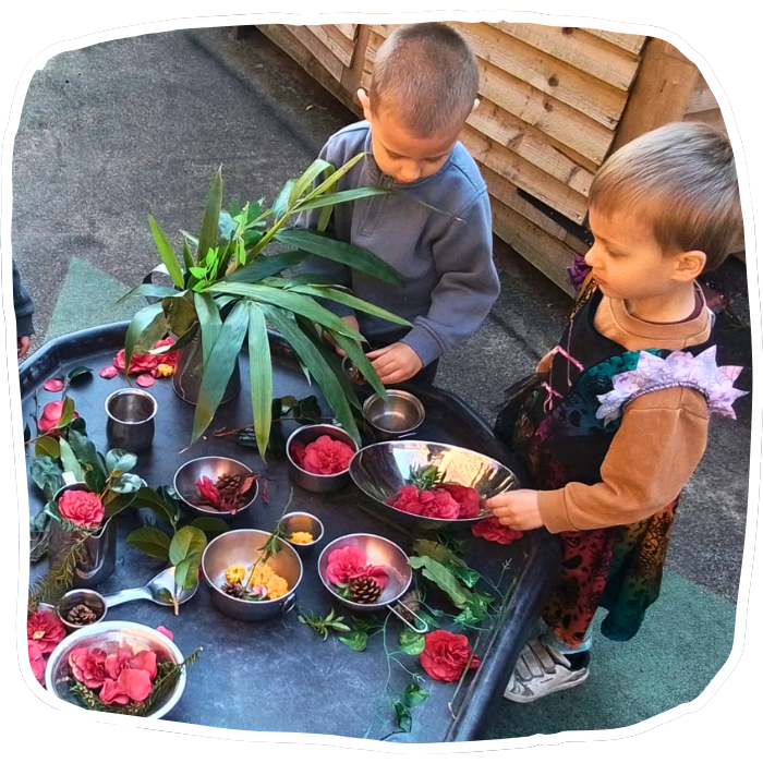 Two young children arrange flowers, leaves, and pinecones in metal bowls on a low outdoor table beside a wooden fence.