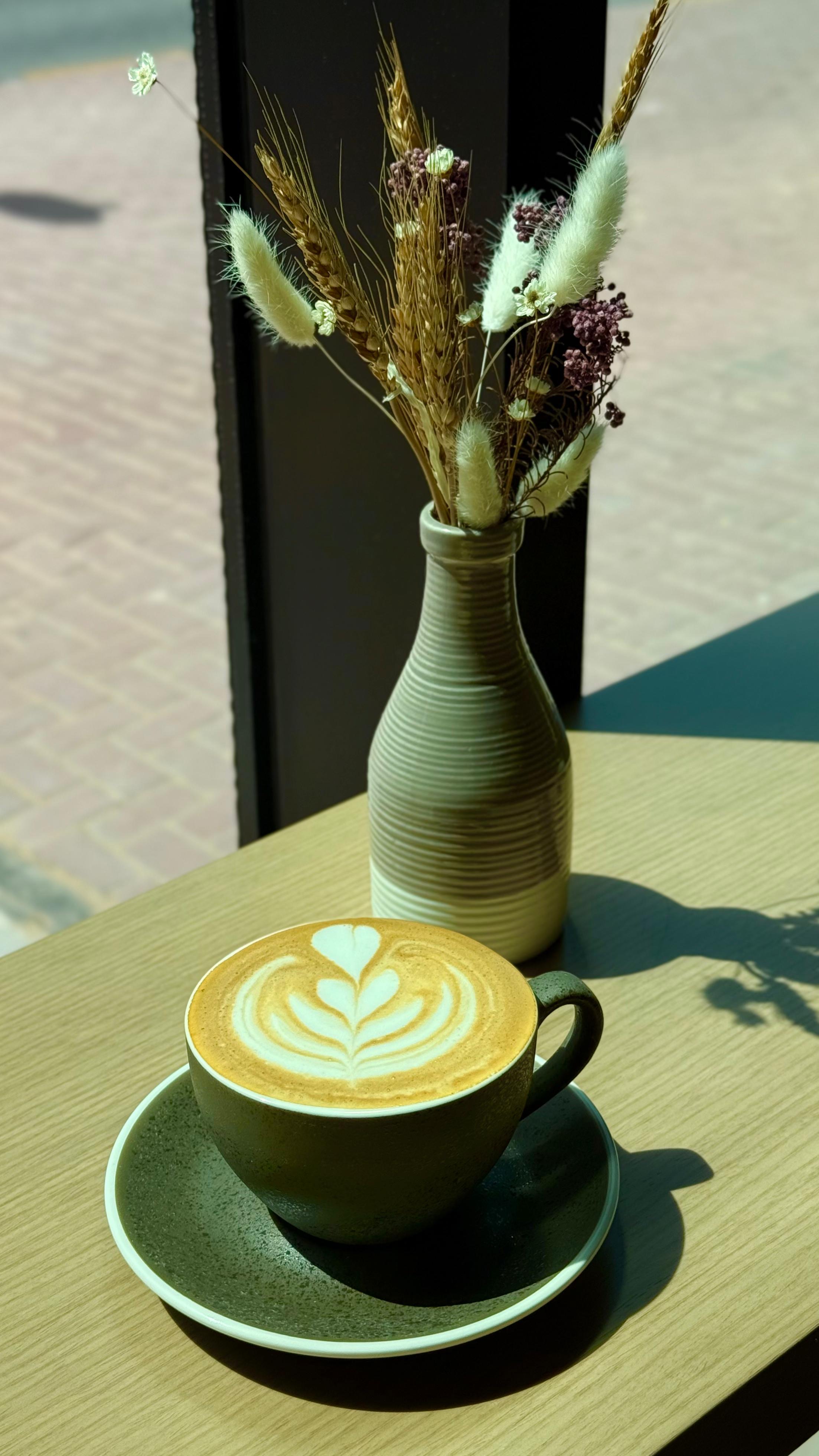 Freshly brewed latte with heart-shaped latte art beside a vase of dried flowers at Adina’s Coffee Shop JVC Dubai