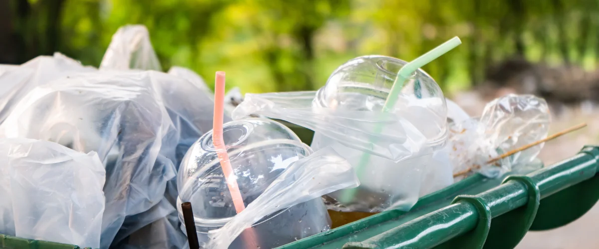 A close-up shot of a green trash bin overflowing with plastic cups, straws, and plastic bags against a blurred green background. A close-up shot of a green trash bin overflowing with plastic cups, straws, and plastic bags against a blurred green background.