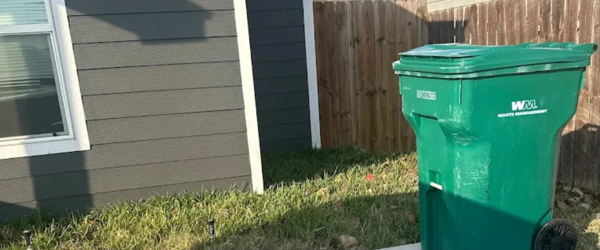 A green Waste Management garbage can sits next to a gray house with a window, against a wooden fence. A green Waste Management garbage can sits next to a gray house with a window, against a wooden fence.