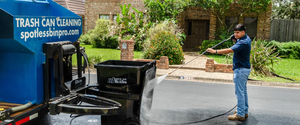 A man in jeans and blue shirt power washes a black trash can on a residential street next to a blue cleaning truck. A man in jeans and blue shirt power washes a black trash can on a residential street next to a blue cleaning truck.