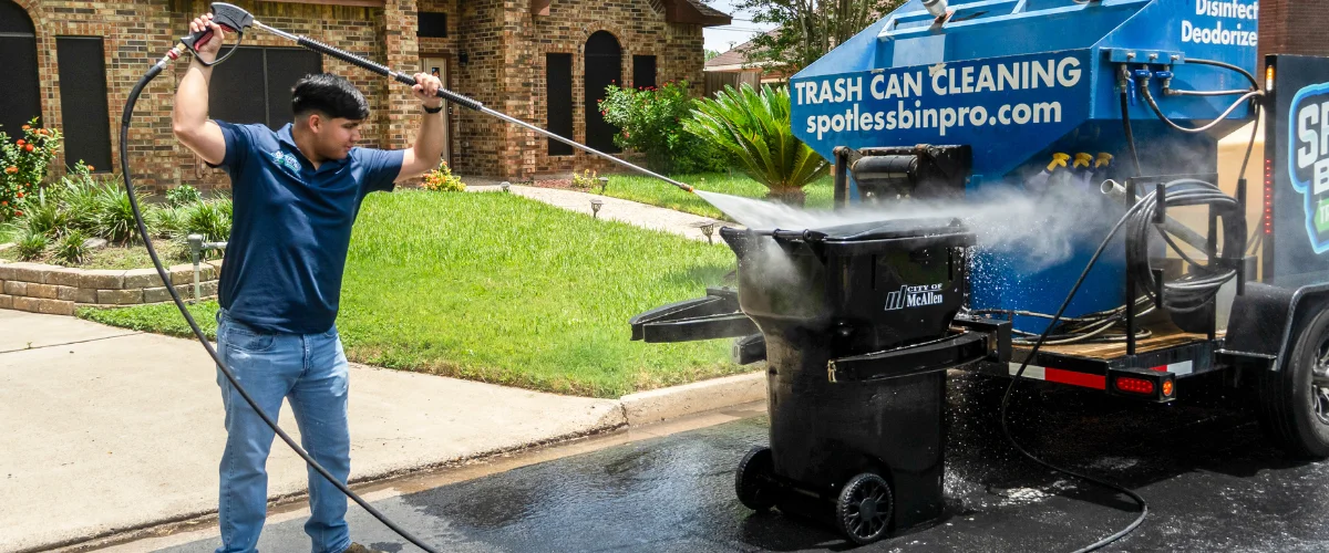 A man in a blue shirt standing on the street, power-washing a black trash can with a long wand. A man in a blue shirt standing on the street, power-washing a black trash can with a long wand.
