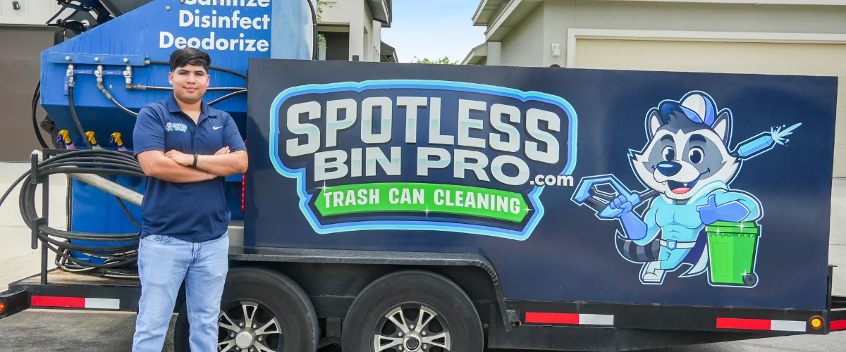 A young man with folded arms stands proudly beside a blue Spotless Bin Pro trash can cleaning trailer.