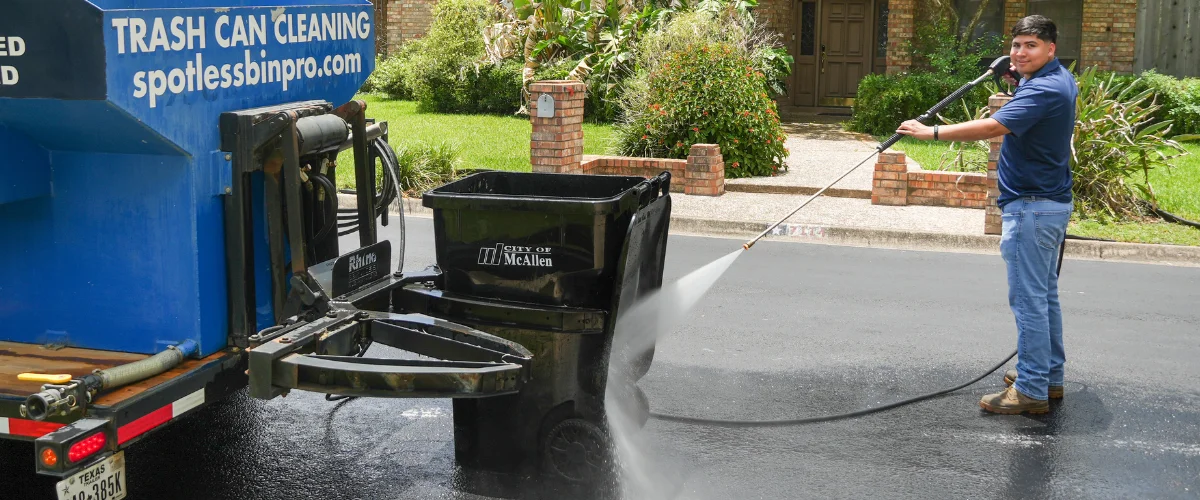 A young man uses a pressure washer to clean a black trash bin next to a blue cleaning service truck on a sunny day.