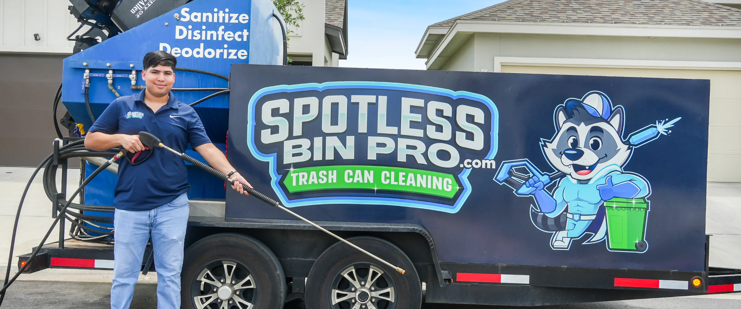 A person in a blue shirt and jeans pressure washing a black City of McAllen garbage bin outdoors.