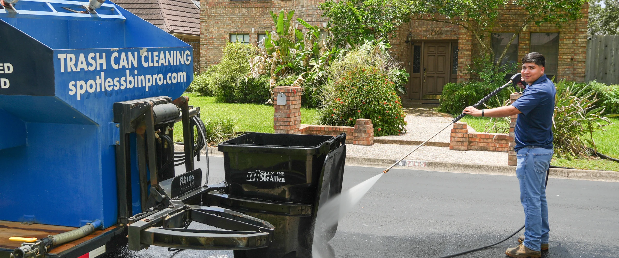 A man in a blue polo shirt and jeans pressure washes a black City of McAllen trash can on a residential street. A man in a blue polo shirt and jeans pressure washes a black City of McAllen trash can on a residential street.