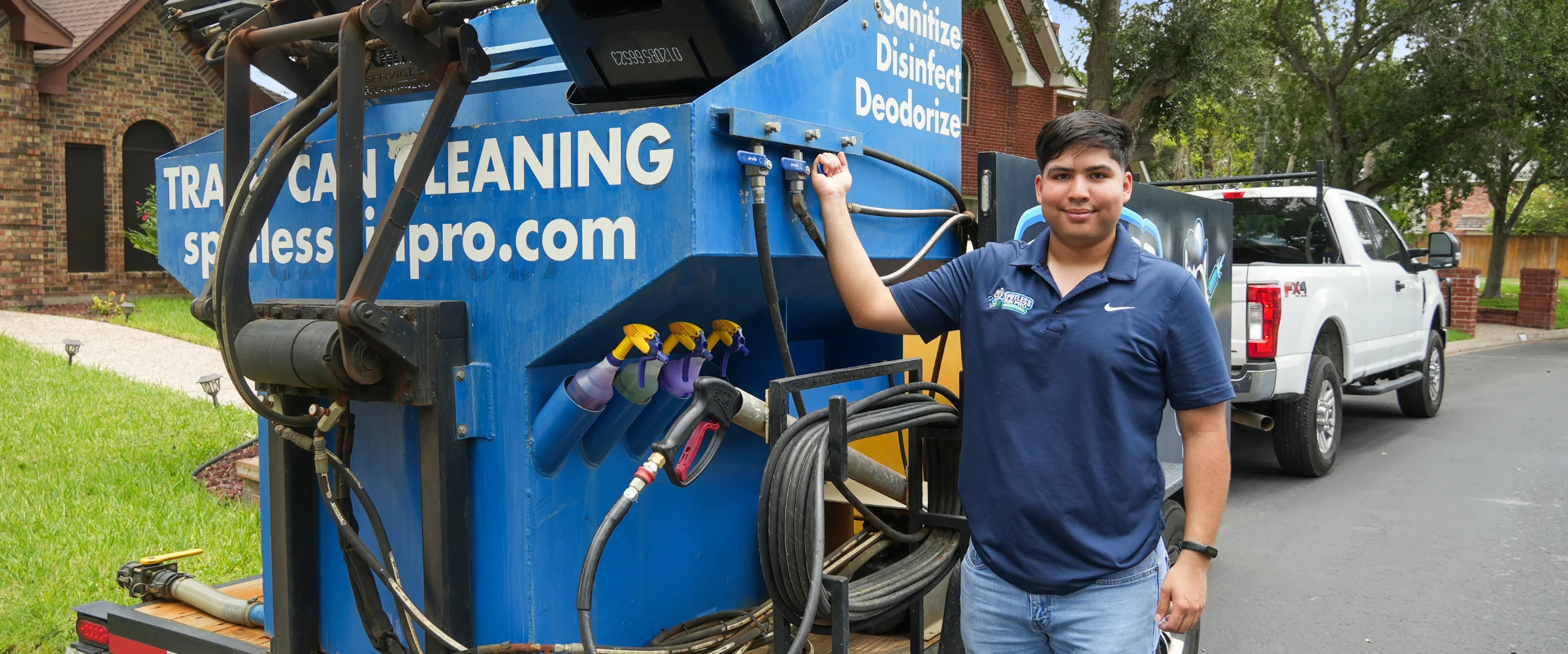 A smiling man in a blue polo shirt stands beside a blue trash can cleaning machine on a trailer. A smiling man in a blue polo shirt stands beside a blue trash can cleaning machine on a trailer.