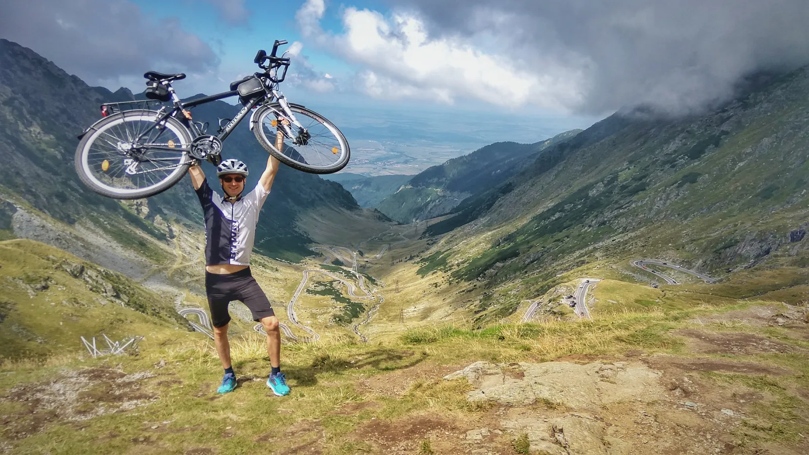 Dr. Konrád ridicând bicicleta deasupra capului, pe un drum de munte
