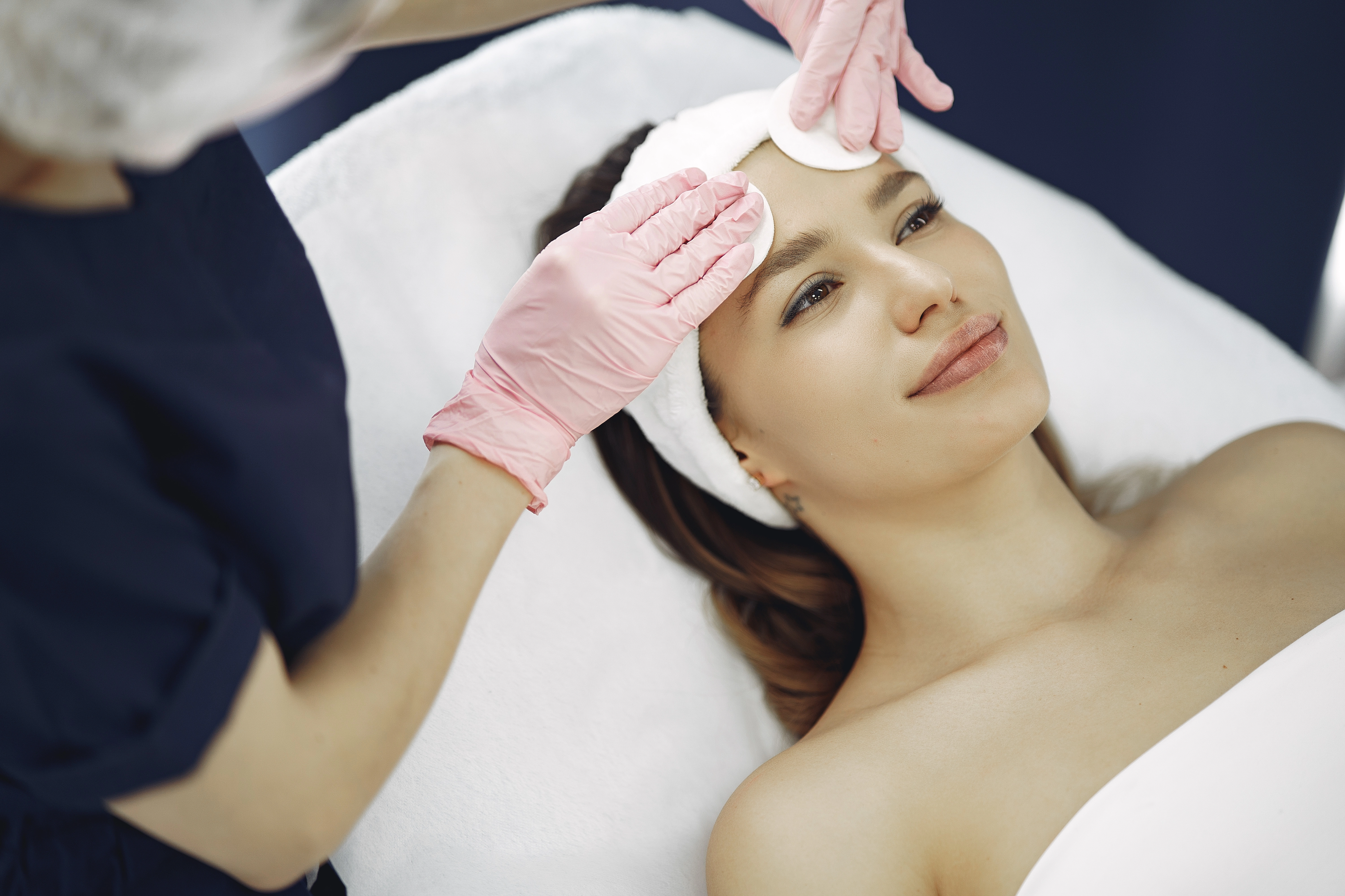 A woman lying comfortably on a treatment bed while a practitioner in gloves gently applies a facial pad to her forehead