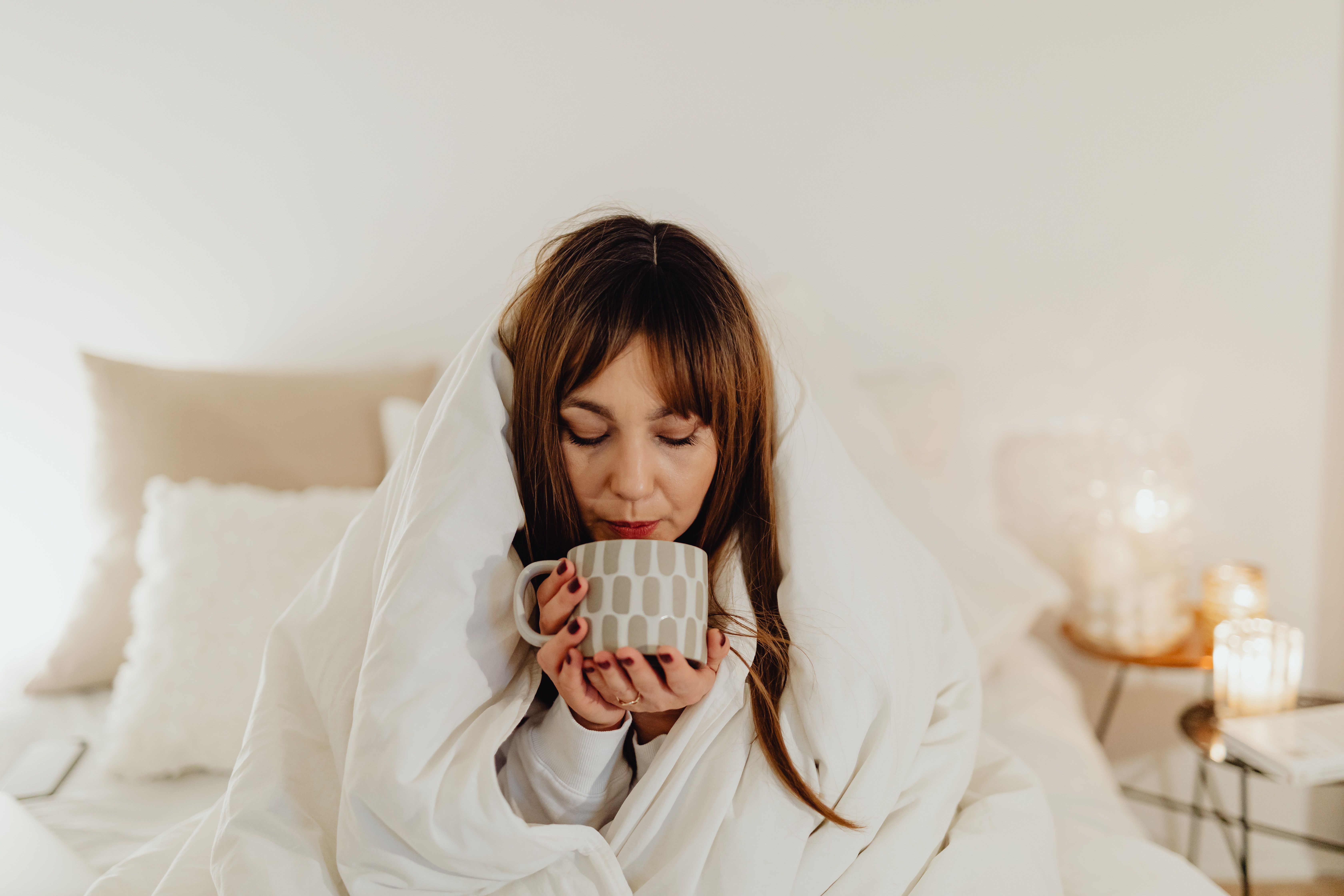 Woman wrapped in a white duvet sipping a warm drink on a cozy fall morning Woman wrapped in a white duvet sipping a warm drink on a cozy fall morning