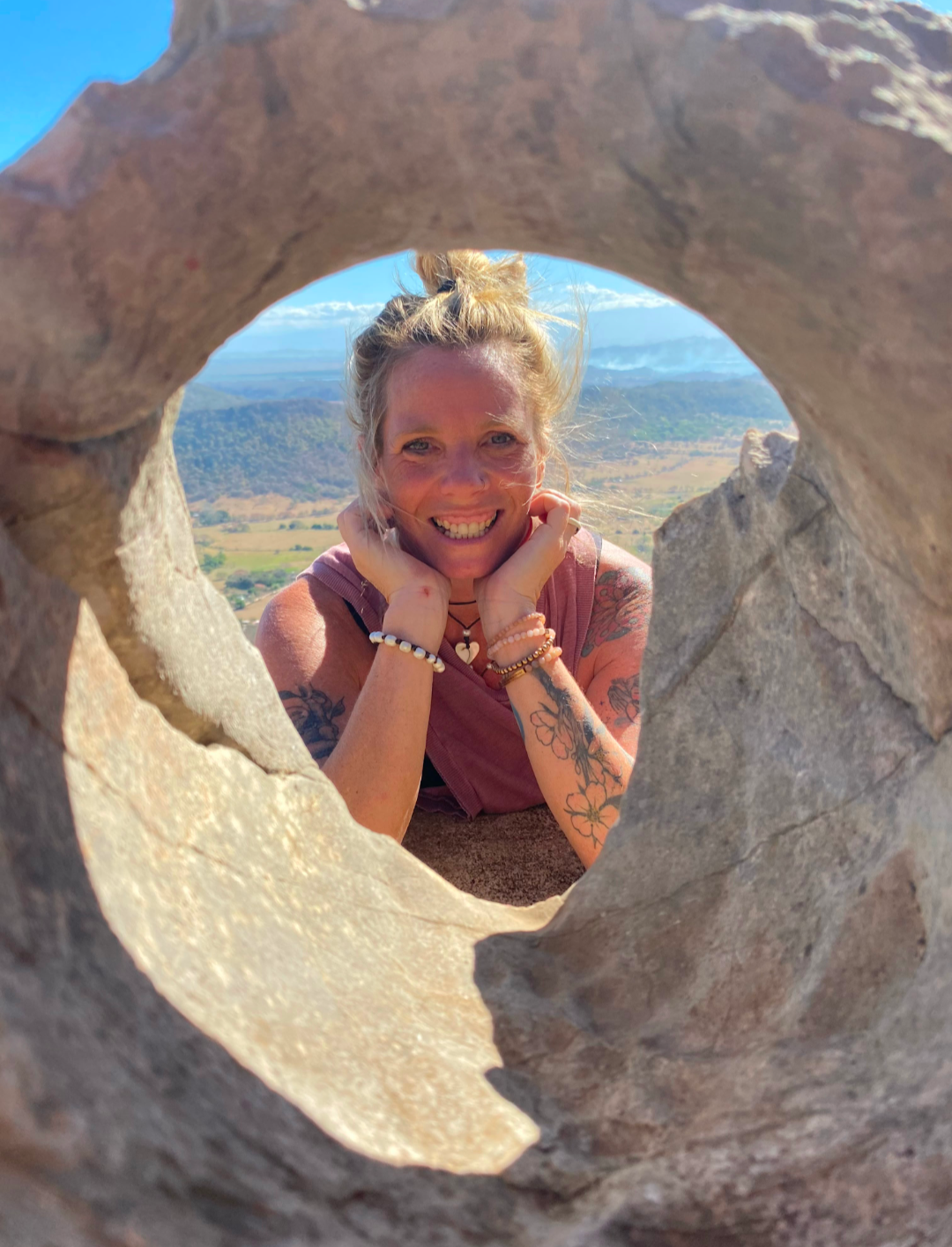 Kari Vincent smiling through an opening in a sandstone rock above a green valley