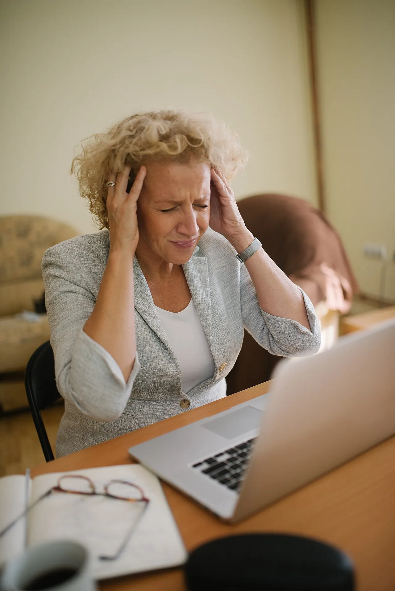 A woman at an office desk holds her head with one hand, appearing to have a headache