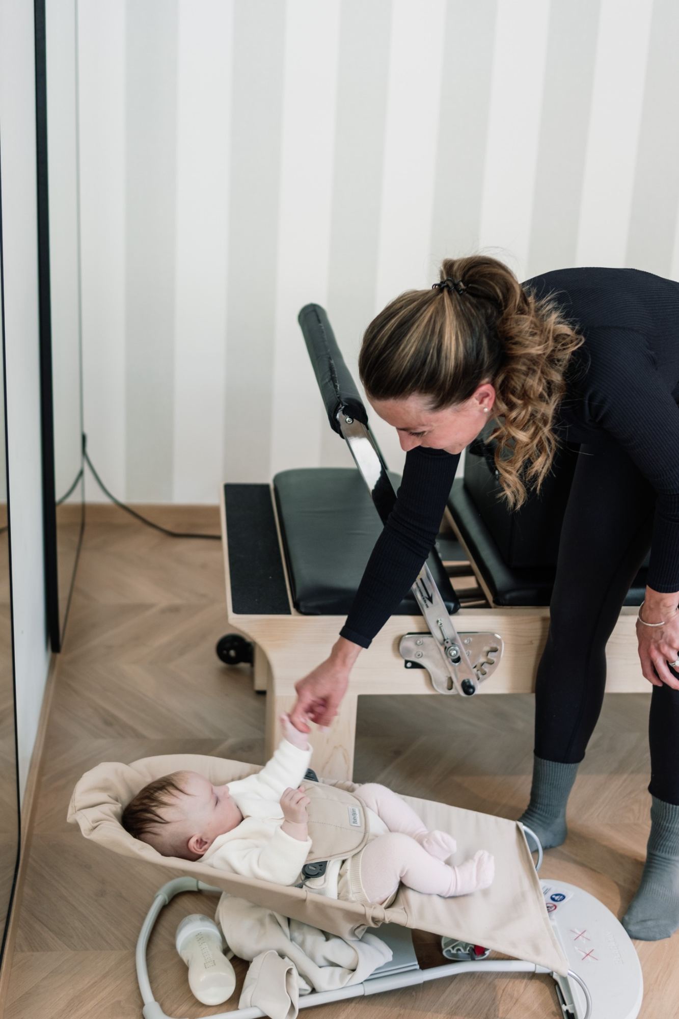 A mom reaching down to her baby in a bouncer next to the Reformer during a Bring Your Baby class