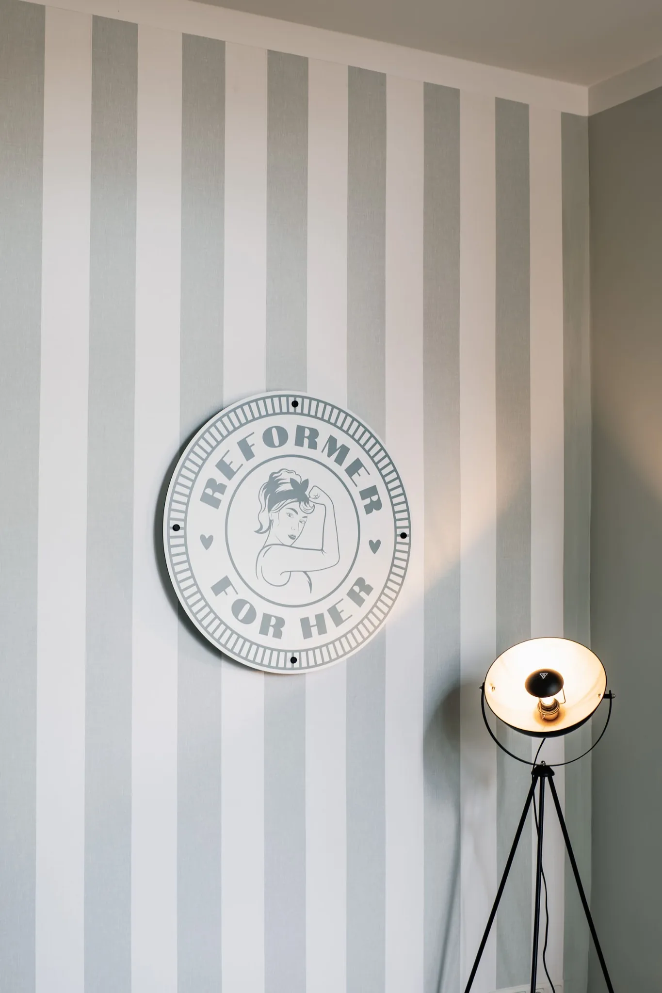 The round Reformer For Her sign on a striped wall next to a vintage spotlight lamp