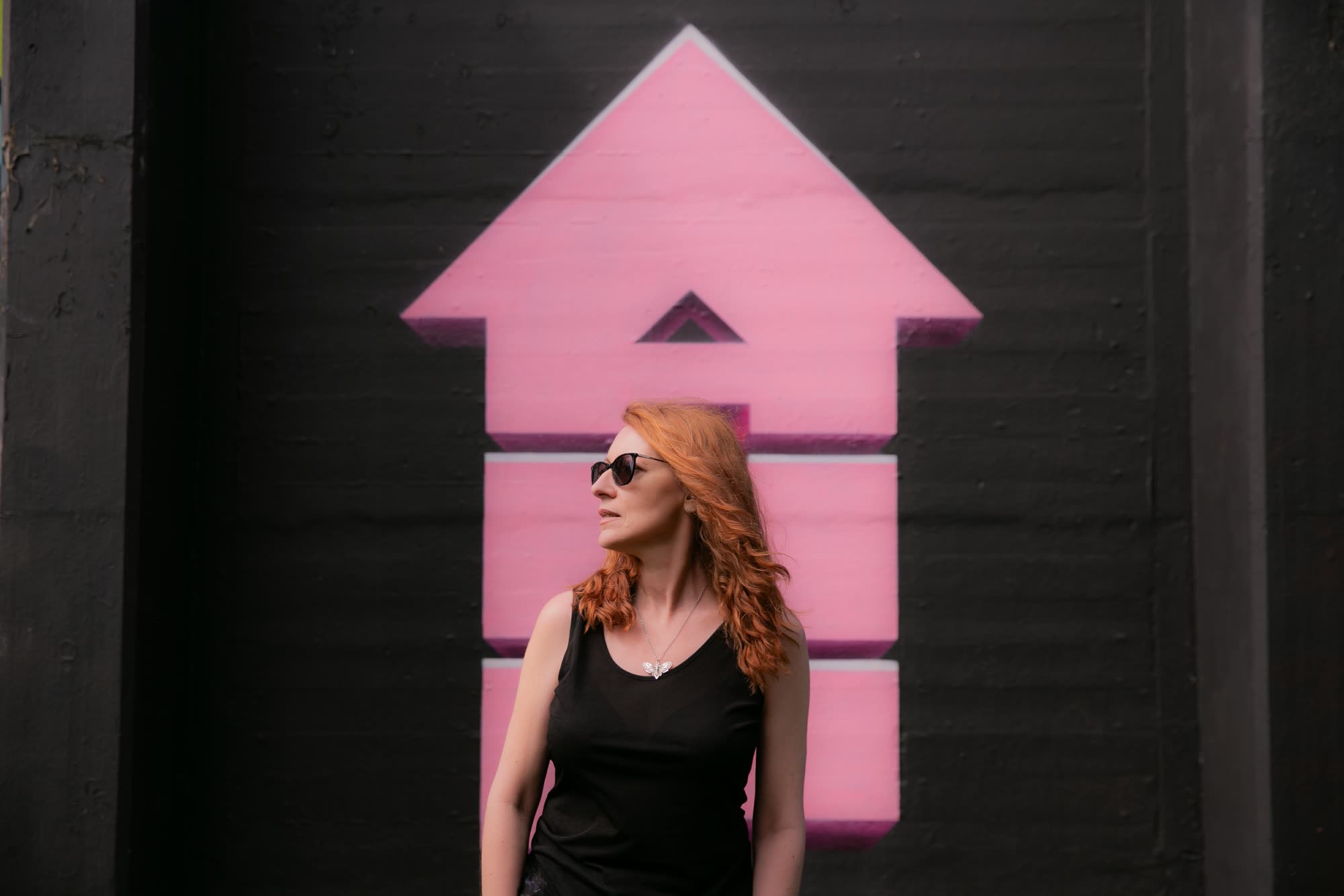Kerry Farrell in a black vest and sunglasses, standing in front of a big pink upward arrow mural on a black wall