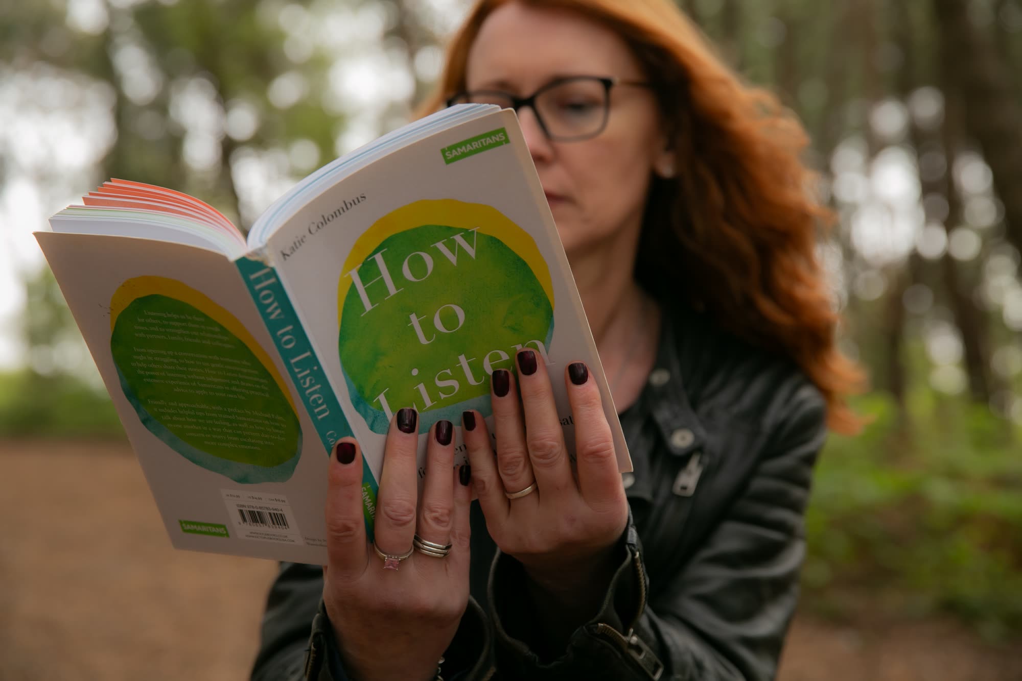 Kerry Farrell in a leather jacket, reading the book How to Listen in a forest