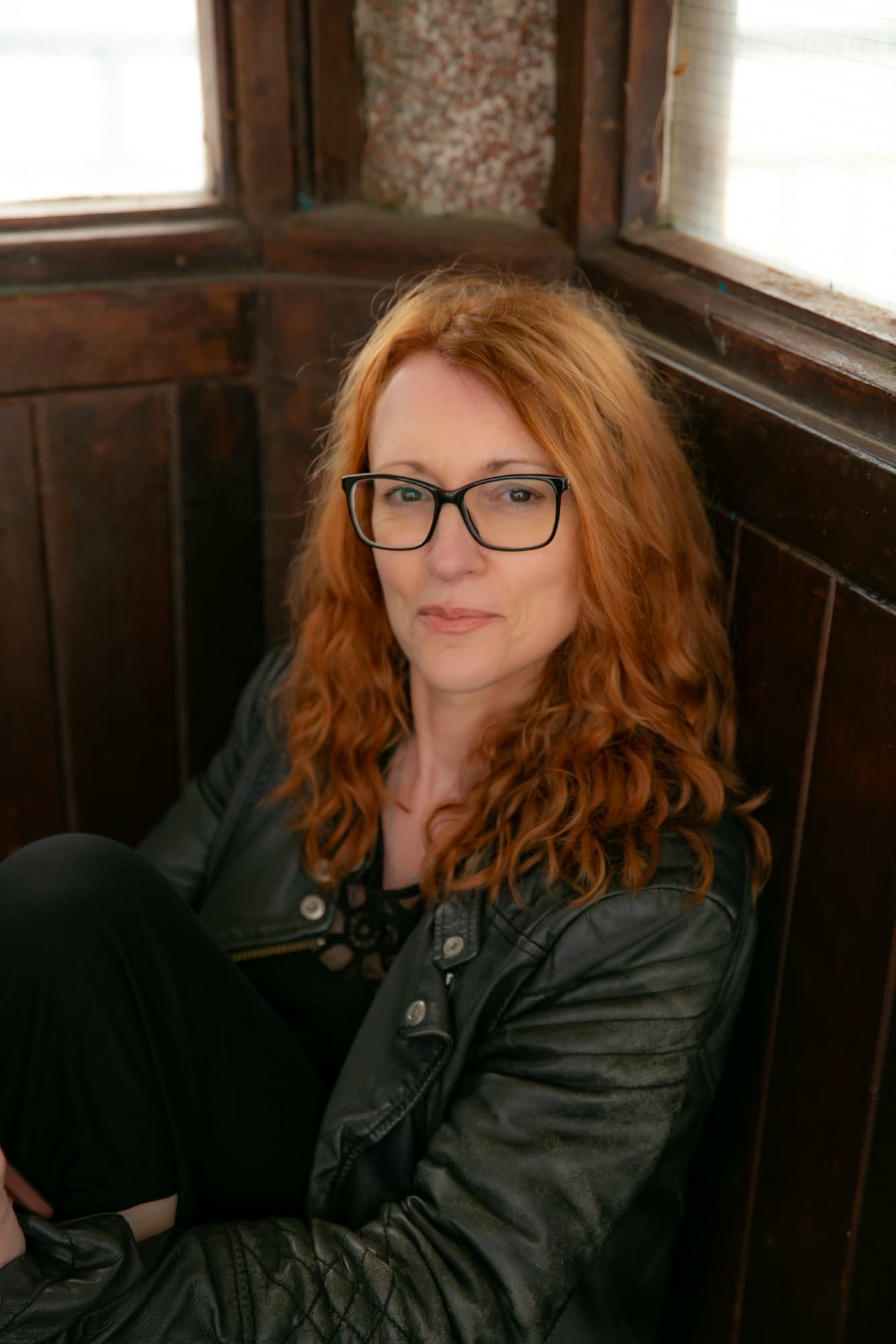 Kerry Farrell in a leather jacket and glasses, sitting in a wooden window seat and looking at the camera