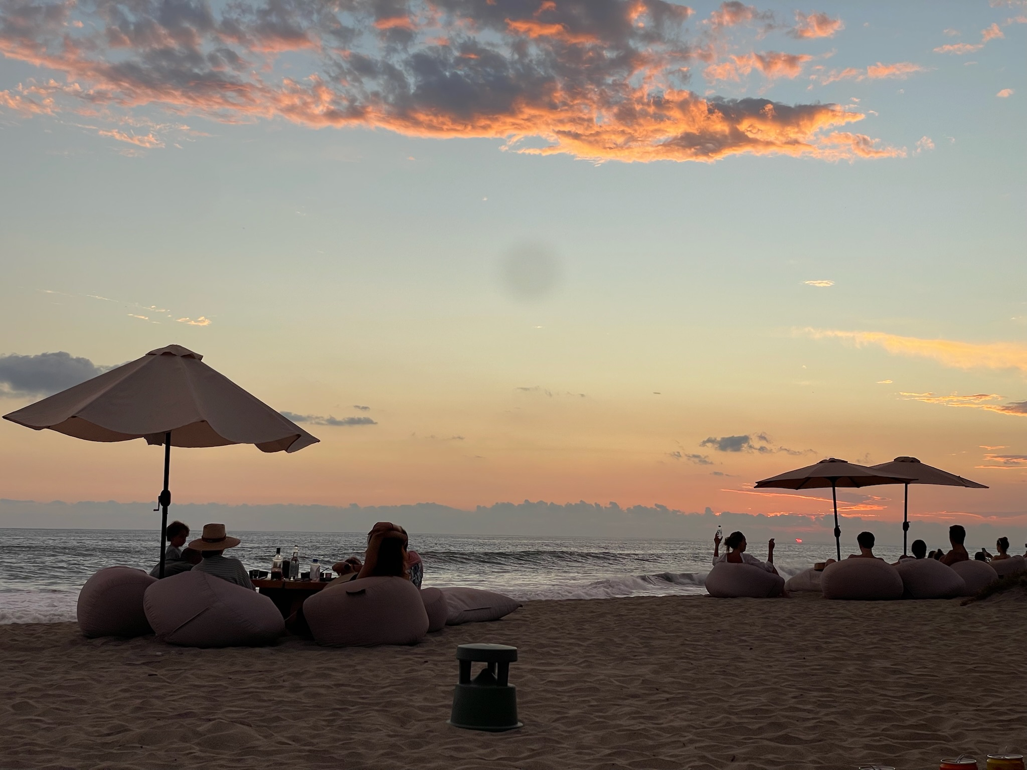 Silhouettes of beachgoers on bean bag chairs under umbrellas watching a pastel sunset over the Pacific Ocean in Puerto Escondido.