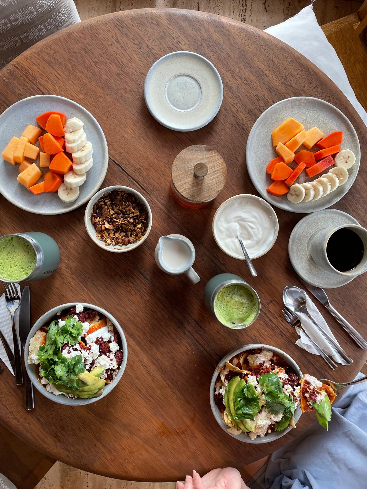 Overhead view of a breakfast spread on a round wood table with fruit plates, granola, yogurt, chilaquiles, and matcha lattes.