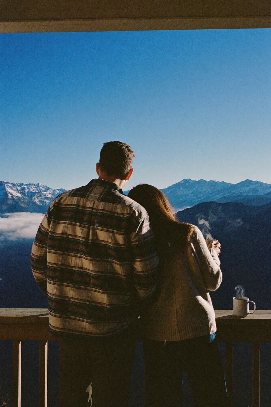 A couple standing close together on a balcony, looking out over snow-capped mountains with morning coffee.