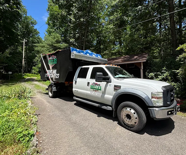 Dirty D's Junk Removal truck delivering a rental dumpster down a wooded residential driveway