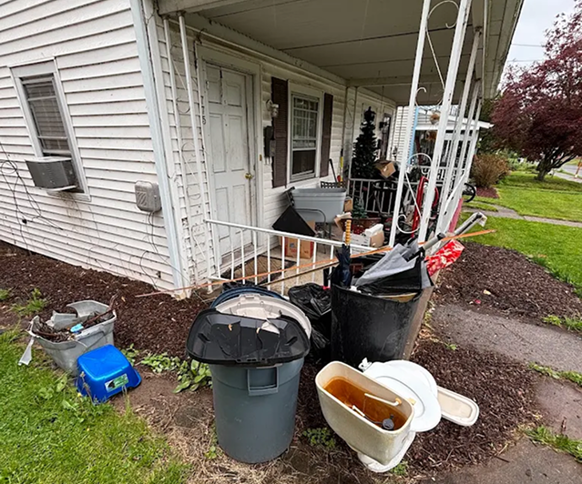 Trash cans, bagged junk, and an old toilet set out by a front porch for curbside junk pickup