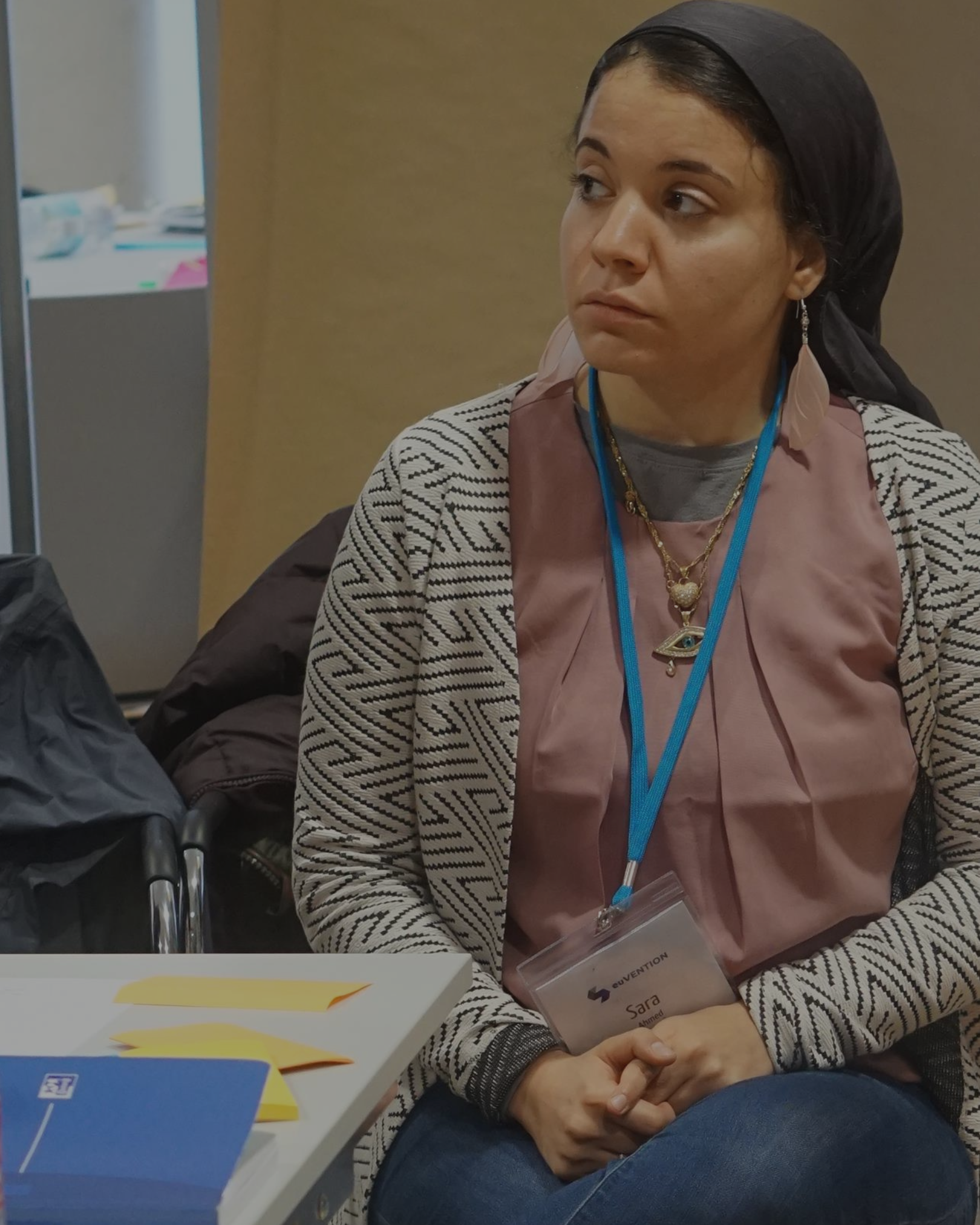 Sara seated at a workshop table, listening quietly with a conference lanyard